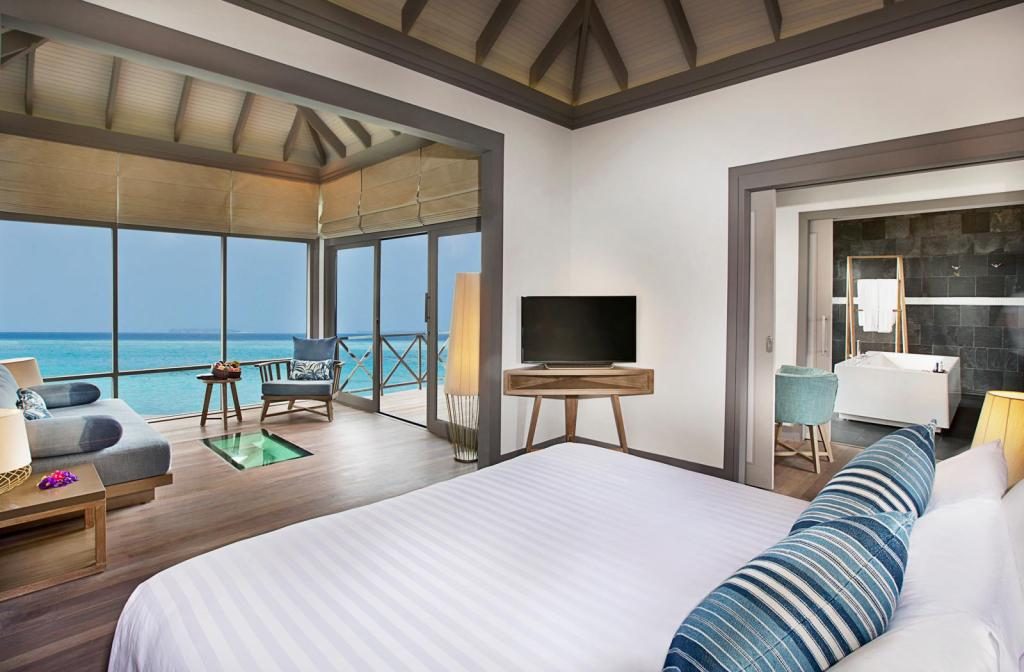 Ja manafaru over water villa room interior with white linen and thatched roof overlooking turquoise lagoon