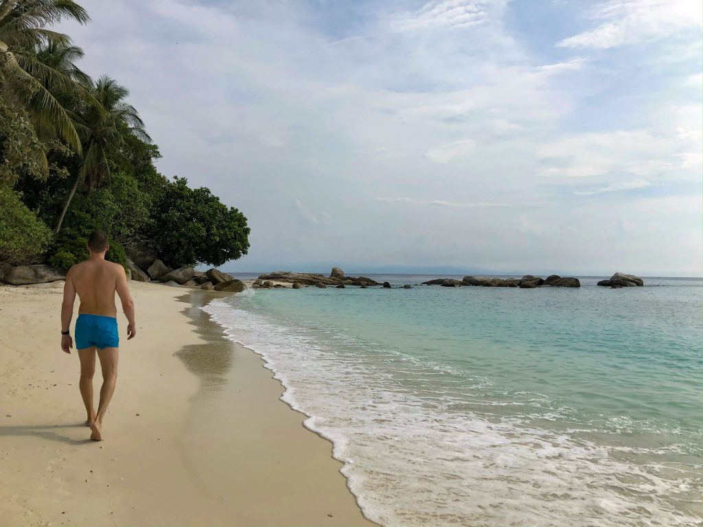 man in blue swim shorts walking along white sand beach towards rocks on lang tengah island