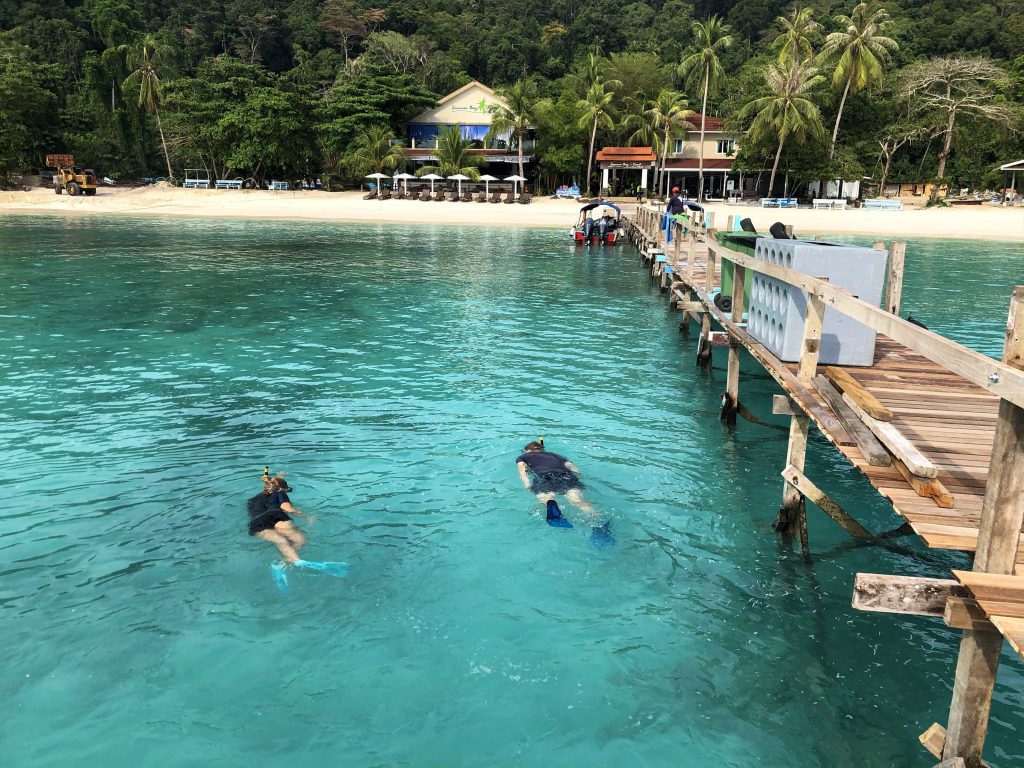 two people snorkeling in crystal turquoise waters next to a boardwalk in lang tengah island