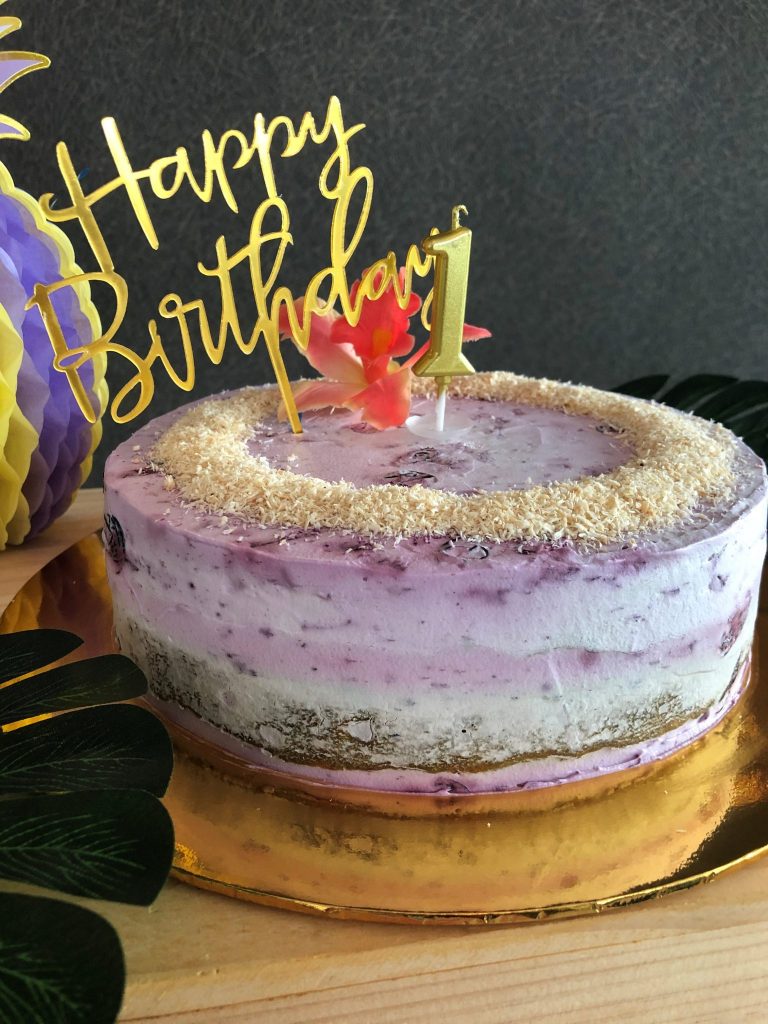 happy 1st first birthday cake topper and 1 candle on a purple frosted baby friendly carrot cake with pineapple and tropical leaves