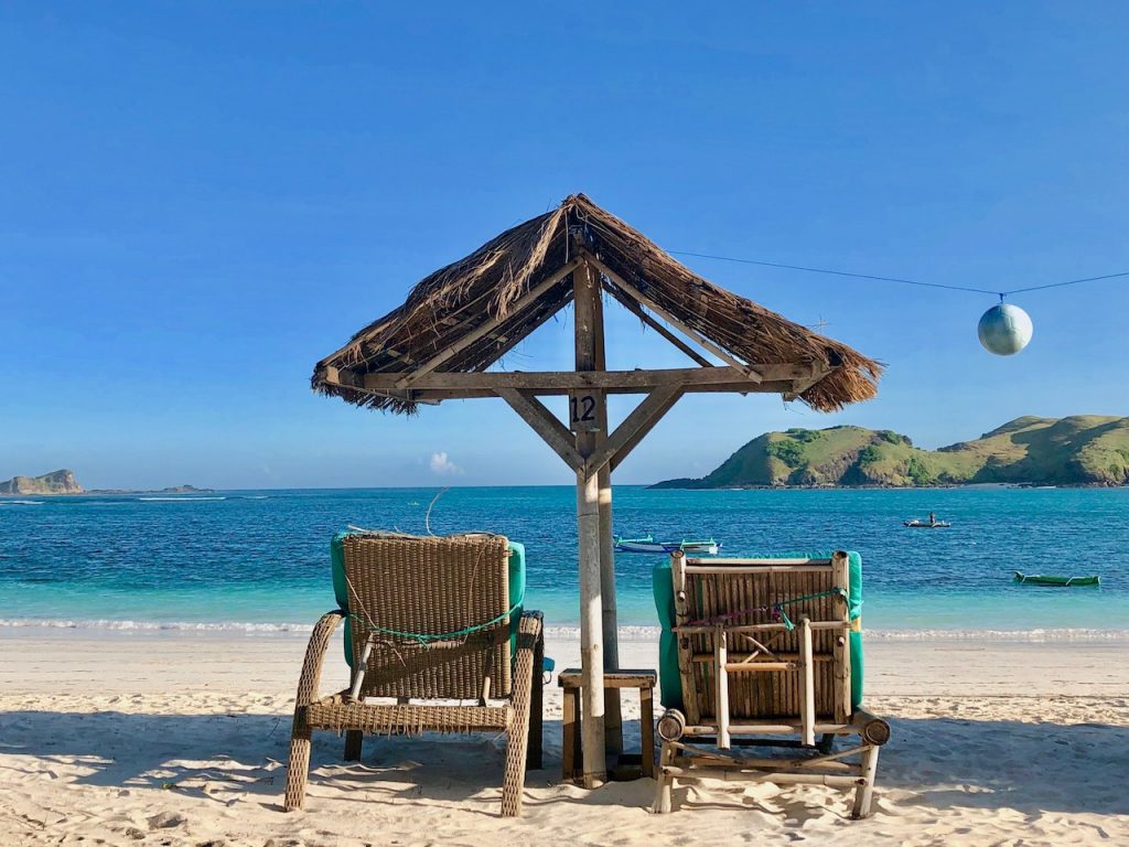 two beach chairs under a thatched canopy on a quiet Tanjung Aan beach in lombok facing blue sea. one of the best secluded beaches around the world