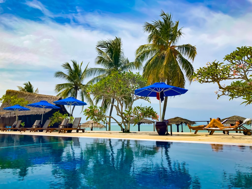 couple tanning by the poolside with swaying palm trees by the beach 