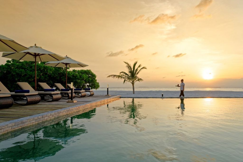 sunset by the beach with reflection of main pool and server with a tray at brennia maldives